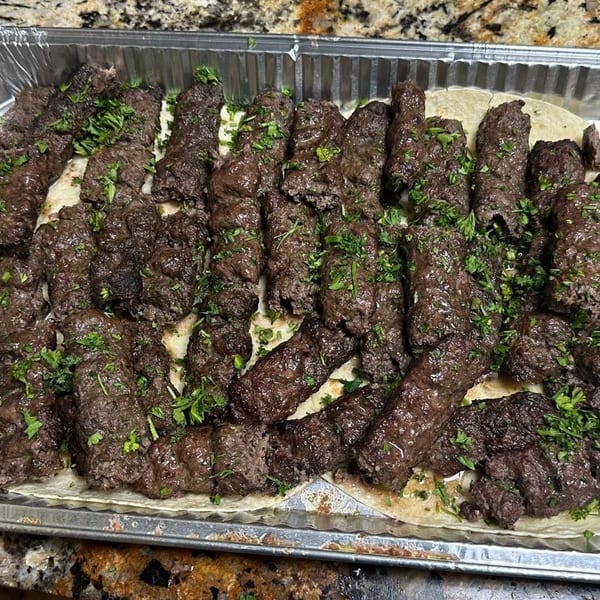 Tray of grilled ground-meat kebabs garnished with chopped parsley, arranged over flatbread in an aluminum catering pan.