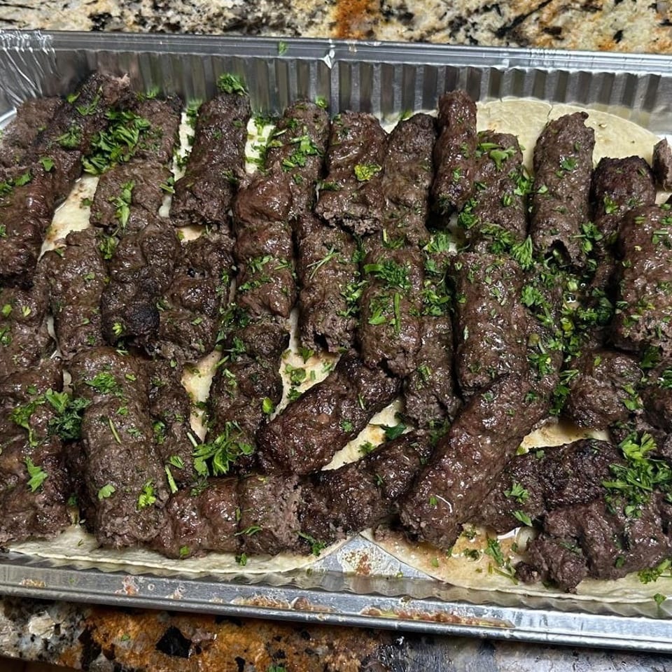 Tray of grilled ground-meat kebabs garnished with chopped parsley, arranged over flatbread in an aluminum catering pan.
