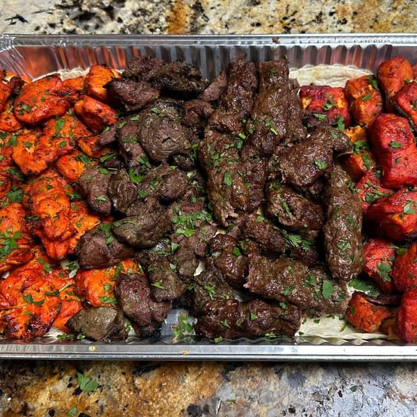 Aluminum tray of grilled chicken, beef kebabs, and colorful roasted vegetables garnished with fresh herbs.