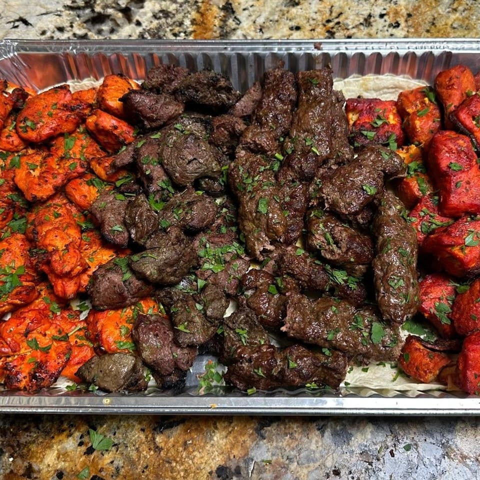 Aluminum tray of grilled chicken, beef kebabs, and colorful roasted vegetables garnished with fresh herbs.