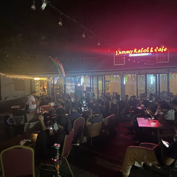 Bustling nighttime outdoor dining area at Jimmy KaloB Restaurant & Cafe, illuminated by string lights and neon signage.