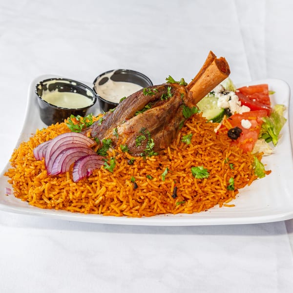 Lamb shank on spiced orange rice with red onions, side salad, and two sauces on a white plate.