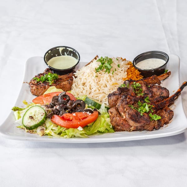 A plate of grilled lamb chops, rice, fresh salad with olives and feta, and two dipping sauces.