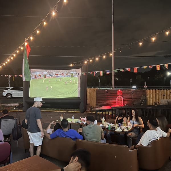 Outdoor restaurant patio with diners watching a soccer match on a large screen beneath string lights and international flags.